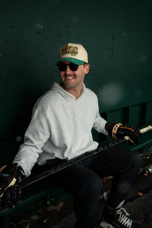 Man wearing Outland Club Co. Dallas City Ice Classic Snapback Hat with sunglasses at a hockey rink, Texas hockey lifestyle photo with hockey stick and gloves
