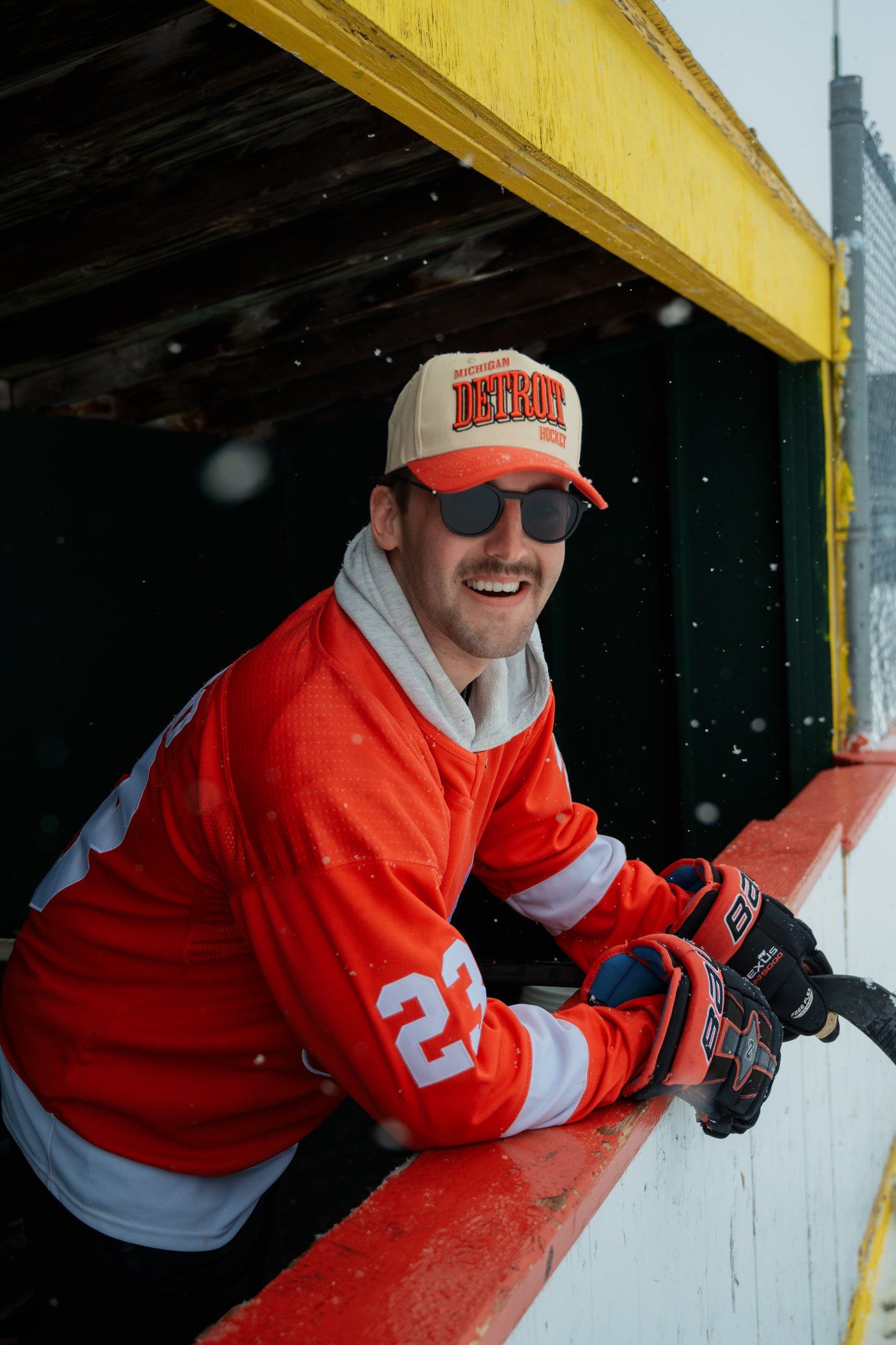 Man wearing Outland Club Co. Detroit City Ice Classic Snapback Hat with sunglasses and hockey jersey at an outdoor rink in winter, hockey lifestyle photo