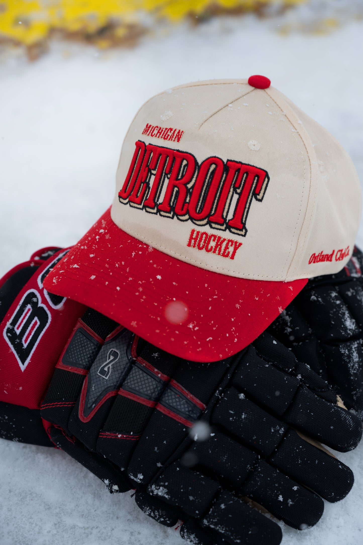 Outland Club Co. Detroit City Ice Classic Snapback Hat resting on hockey gloves in snow, cream crown with red brim and DETROIT HOCKEY embroidery close-up